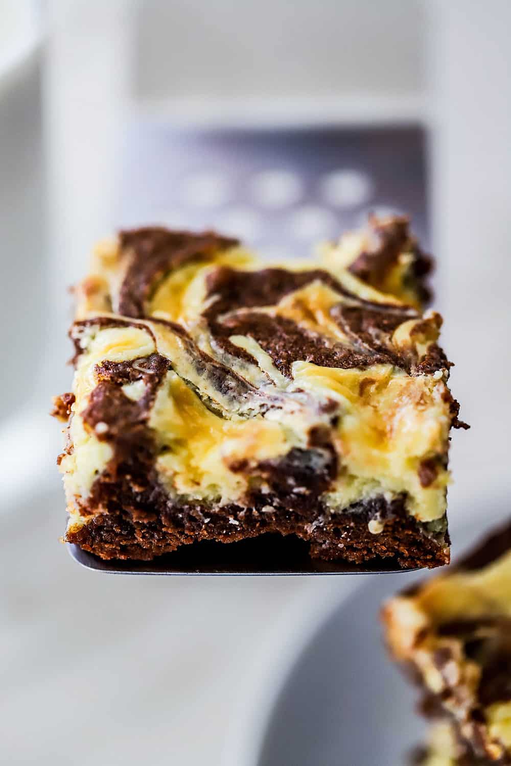 A chocolate and cheese cake brownies being held up on a metal spatula.