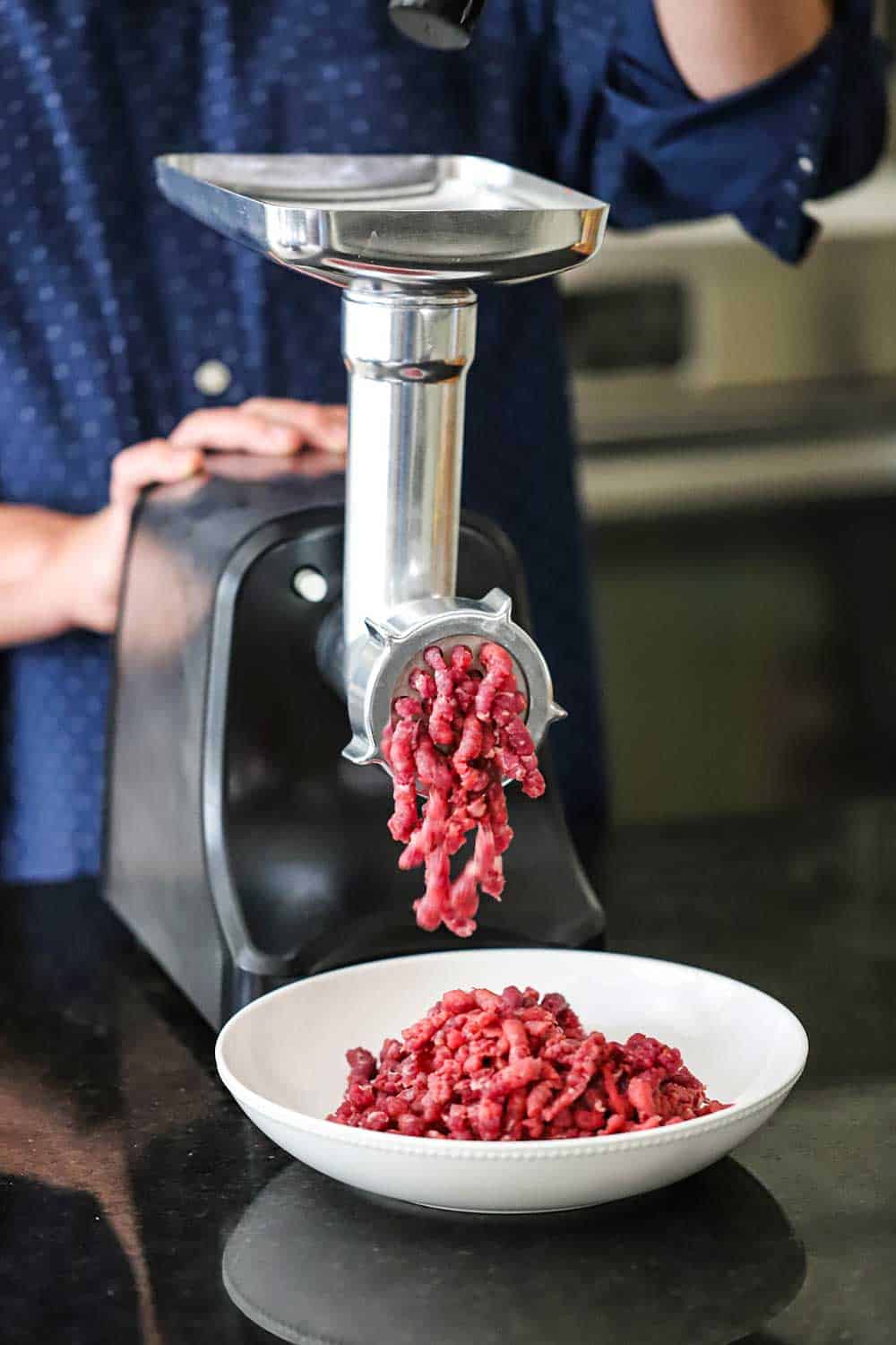 A person standing behind a free-standing meat grinder with ground beef coming out of the grinder and falling into a white bowl on the counter.