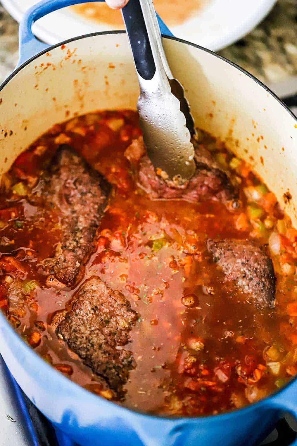 A pair of tongs nestling a cube steak into a large blue Dutch oven filled with other steaks and a chunky tomato sauce.