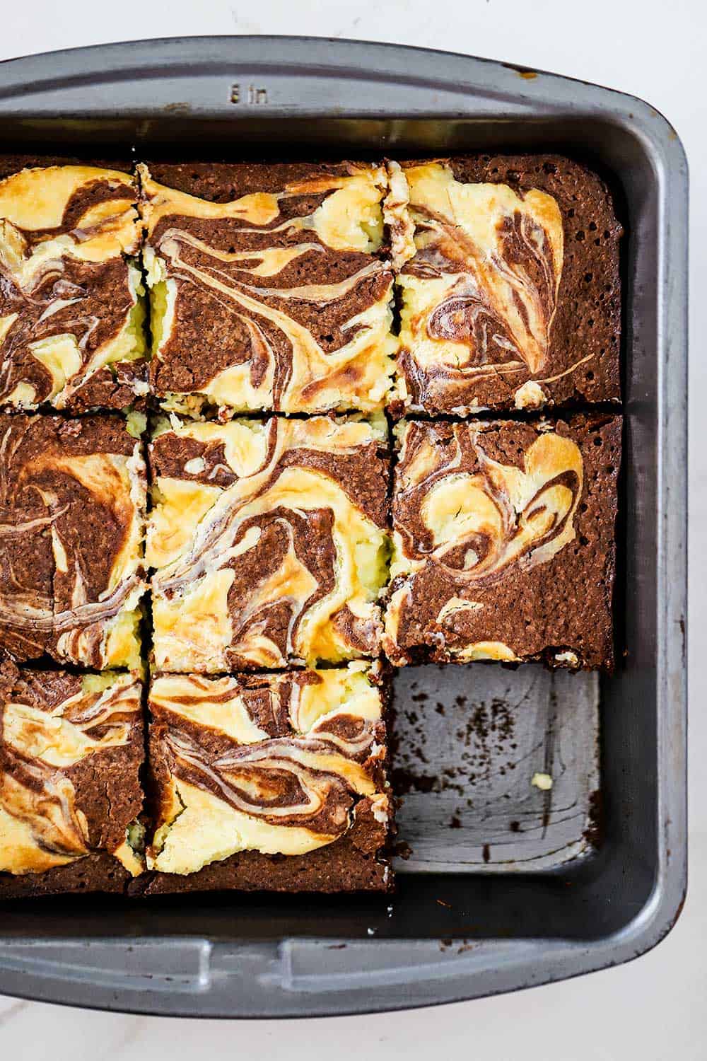 A metal pan filled with chocolate and cheesecake brownies that have been cut into squares and one brownie is missing.