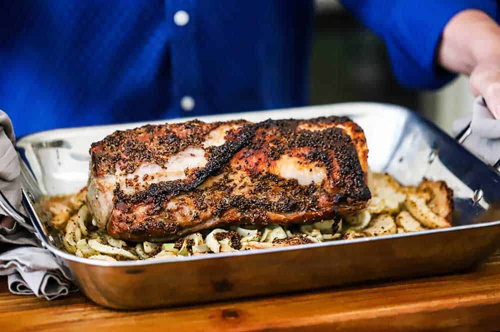 A person holding a large steel roasting pan filled with a roasted pork loin sitting on top of roasted apples and fennel.