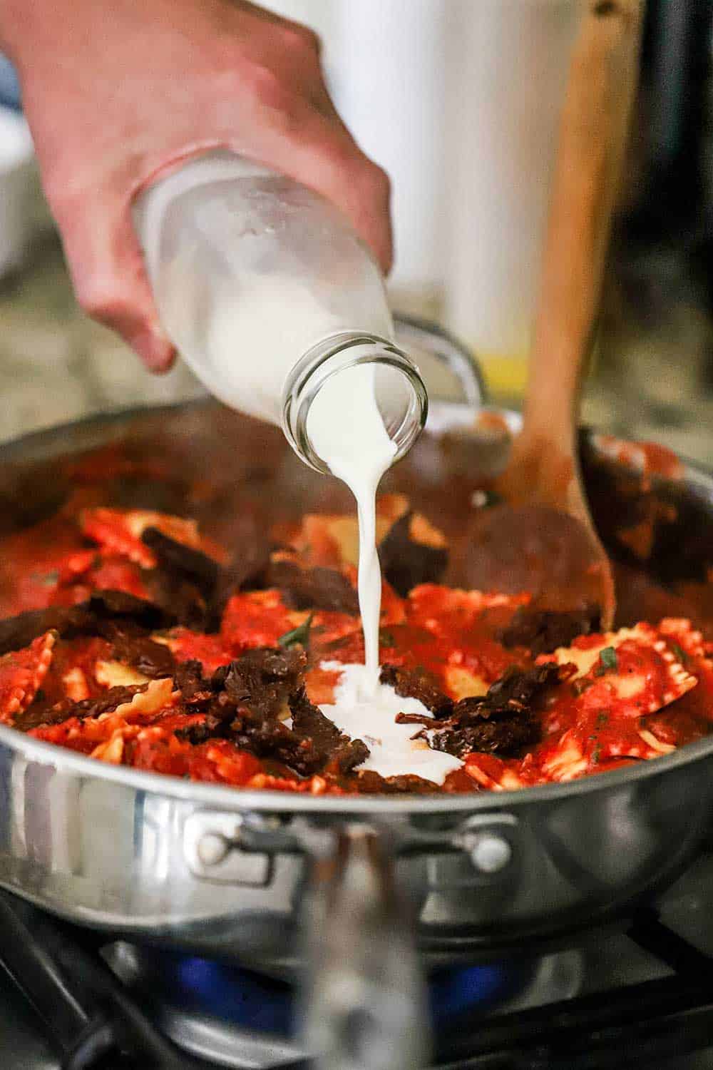 A person pouring cream from a small milk bottle into a skillet filled with ravioli, sun-dried tomatoes, and a tomato sauce. 