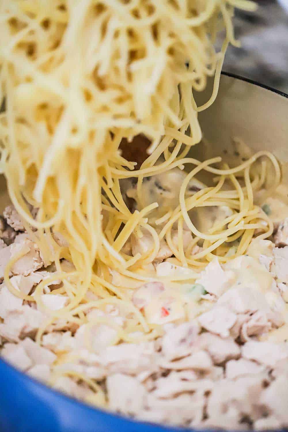 Cooked spaghetti being transferred into a Dutch oven filled with cubed cook chicken and a tomato cheese sauce for chicken spaghetti. 