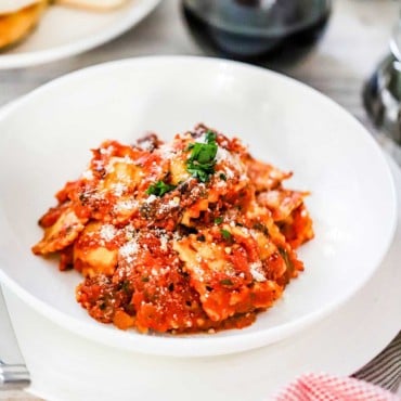 An individual white pasta bowl filled with ravioli and a tomato sauce next to a stemless wine glass filled with red wine and a plate of garlic bread nearby.