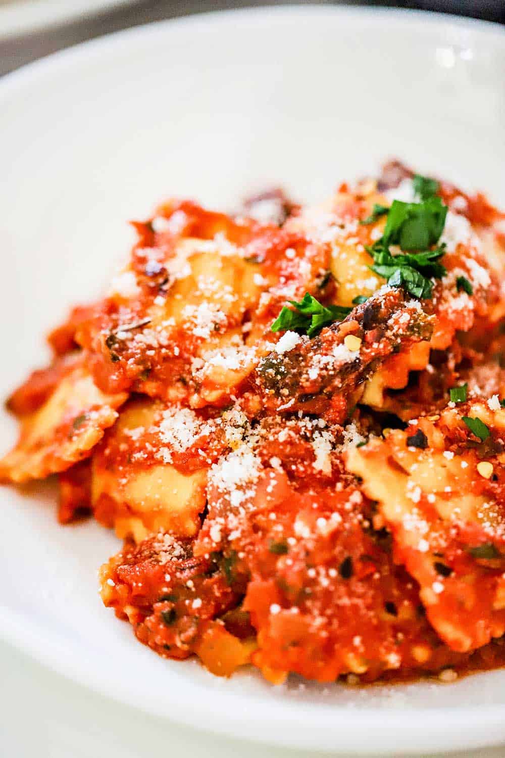 A white individual pasta bowl filled with easy ravioli with creamy tomato-basil sauce with chopped parsley sprinkled on top. 