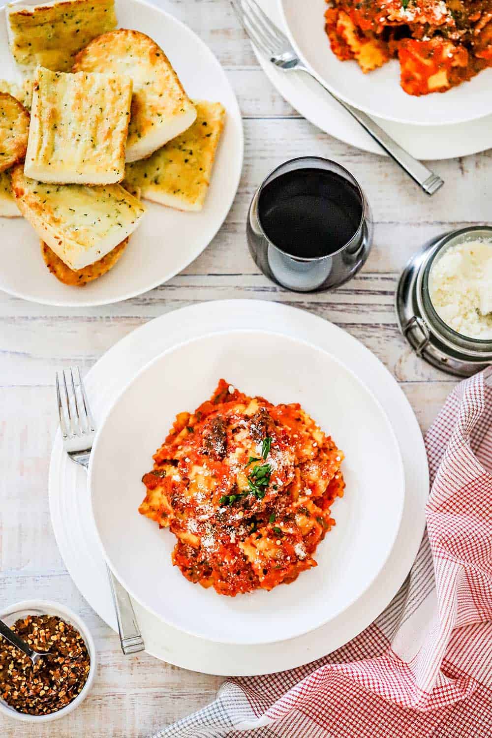 An individual round pasta bowl on a plate filled with easy ravioli and creamy basil-tomato sauce next to a glass of red wine and a plate of sliced garlic bread. 