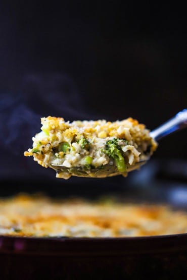 A large silver spoon lifting up a steaming helping of broccoli casserole over a dish of the same.