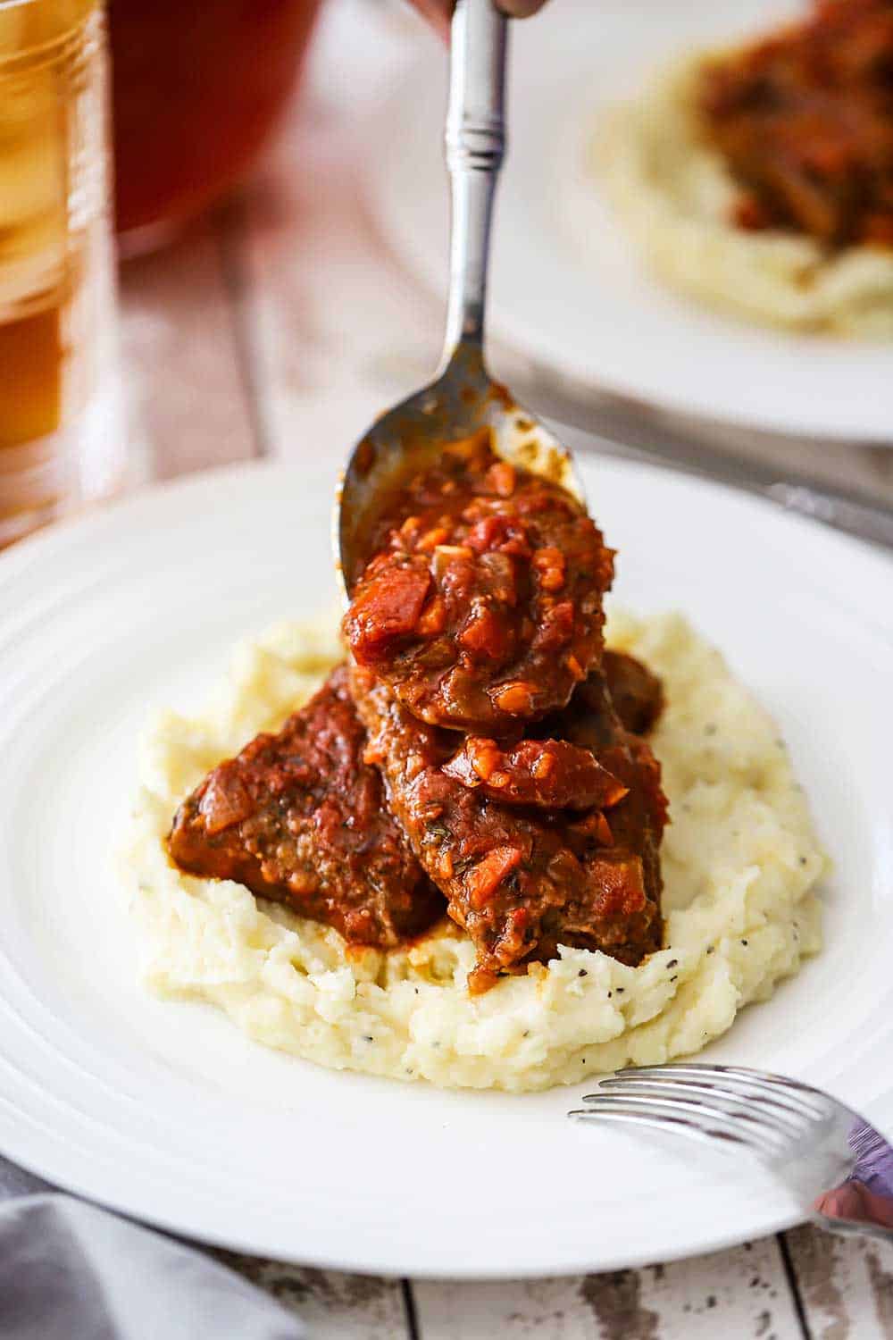 A person using a large silver spoon to pour Swiss steak sauce over two piece of meat sitting on a bed of mashed potatoes on a white dinner plate.