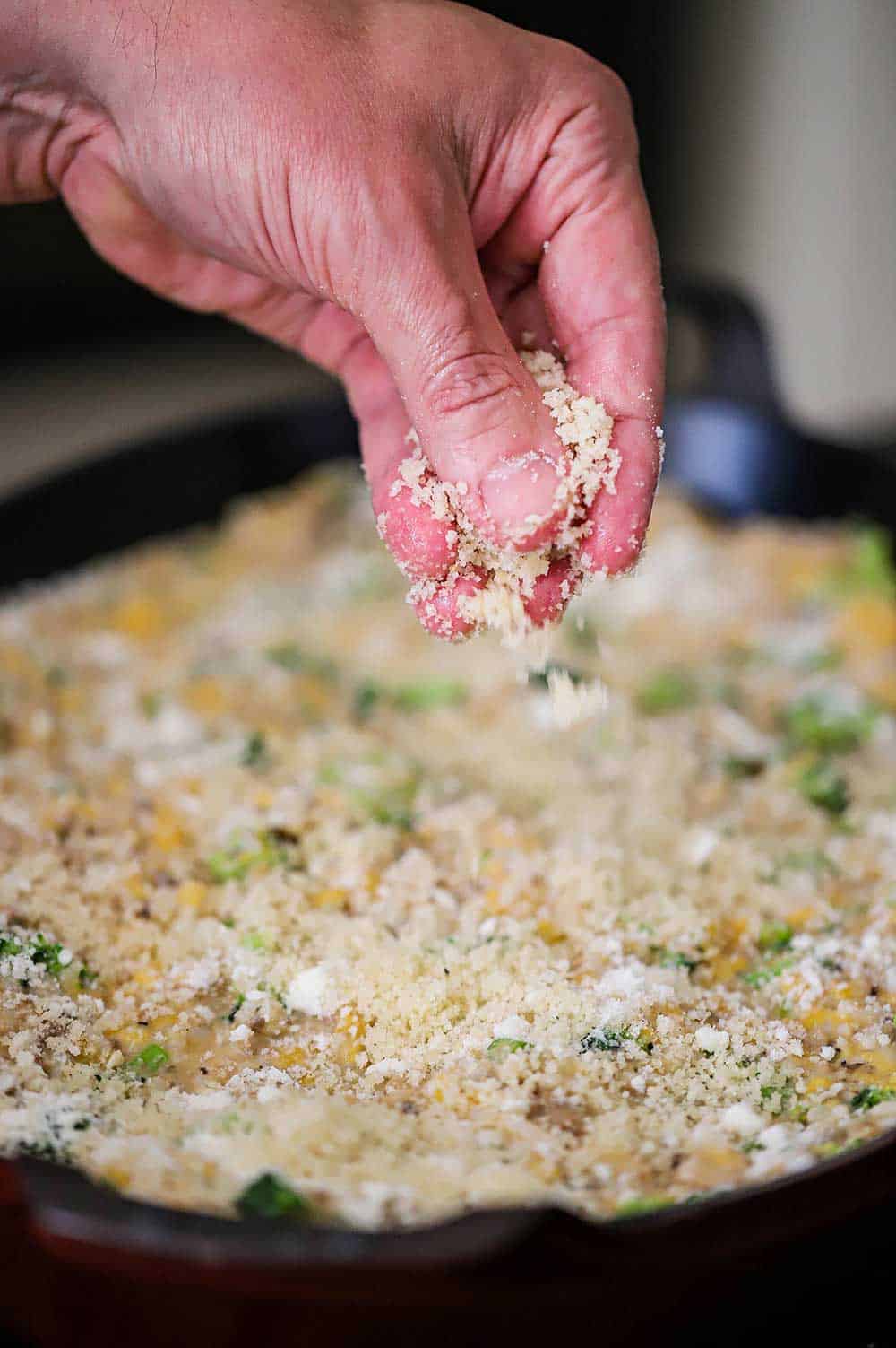 A person sprinkling bread crumbs over the top of an uncooked broccoli casserole in a large oval baking dish.