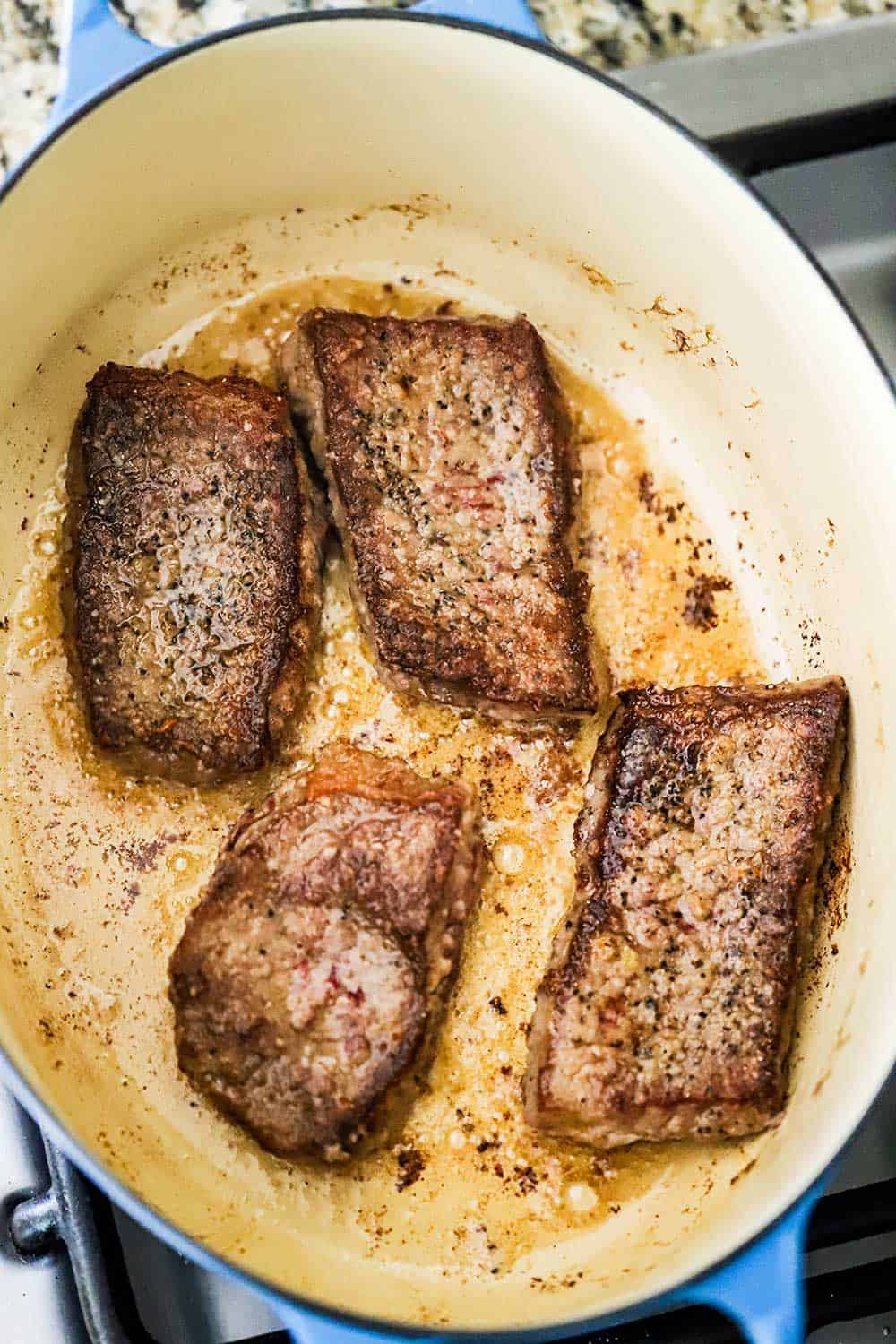 A large oval Dutch oven filled with 4 steaks that have been seared and browned in bacon grease.