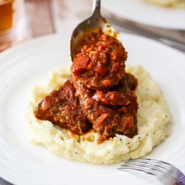 A person using a large silver spoon to pour Swiss steak sauce over two piece of meat sitting on a bed of mashed potatoes on a white dinner plate.