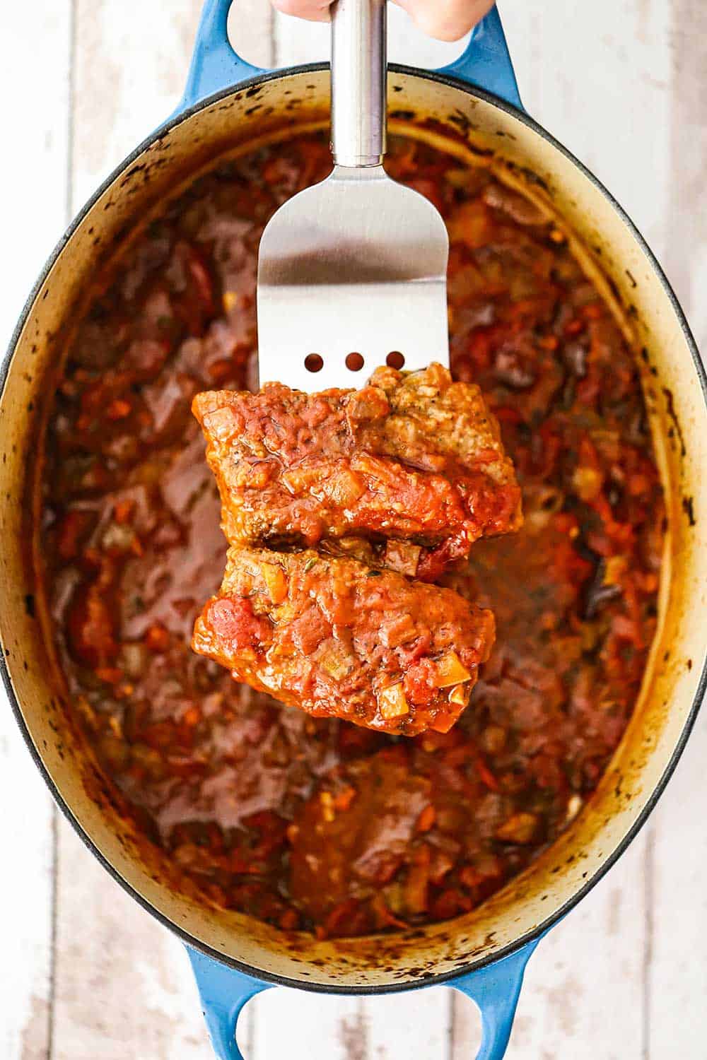 A metal spatula holding up two steak fillets covered in a red tomato sauce directly over an oval Dutch oven filled with Swiss steak.