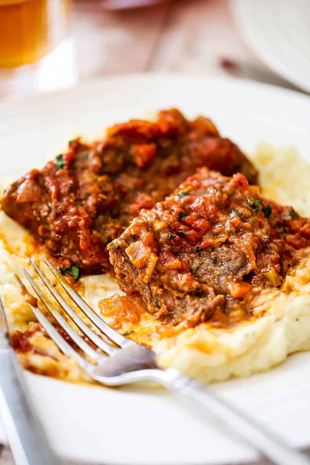 A white dinner plate filled with mashed potatoes topped with a couple of Swiss steaks with a piece missing all next to a sliver fork.