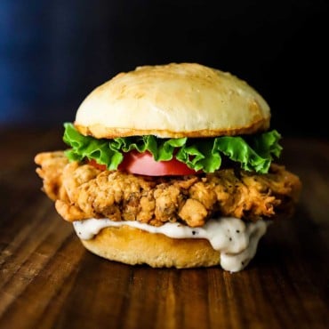 A chicken fried steak sandwich topped with sliced tomato and lettuce sitting on a wooden cutting board.