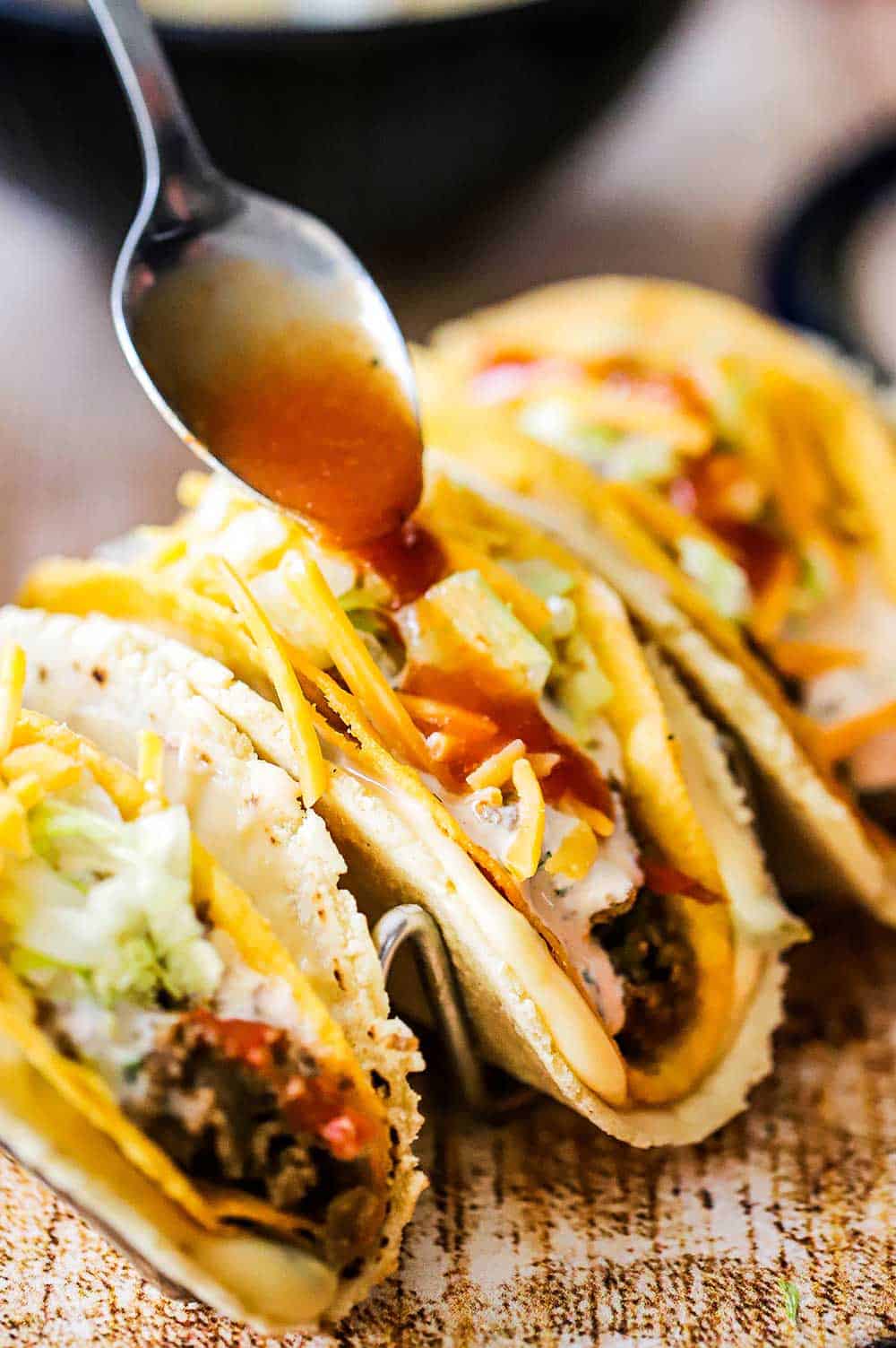 A spoon pouring taco sauce over the top of three stuffed tacos sitting on a metal taco holder.