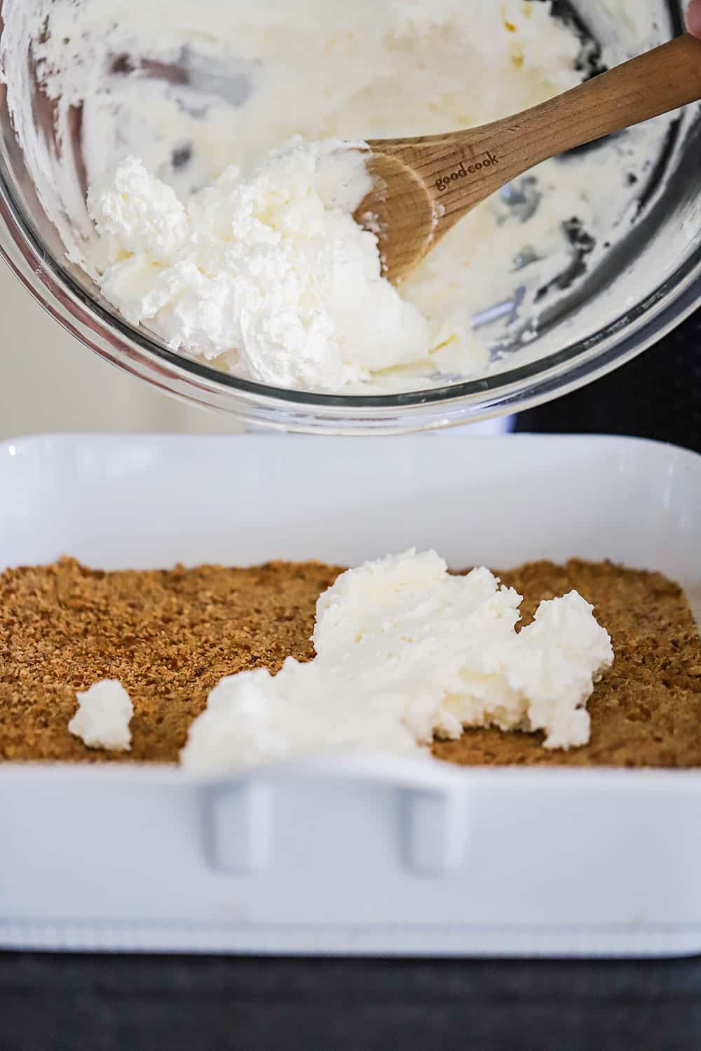 A large glass bowl filled with cream cheese filling being held over a white dish filled with a pretzel crust. 