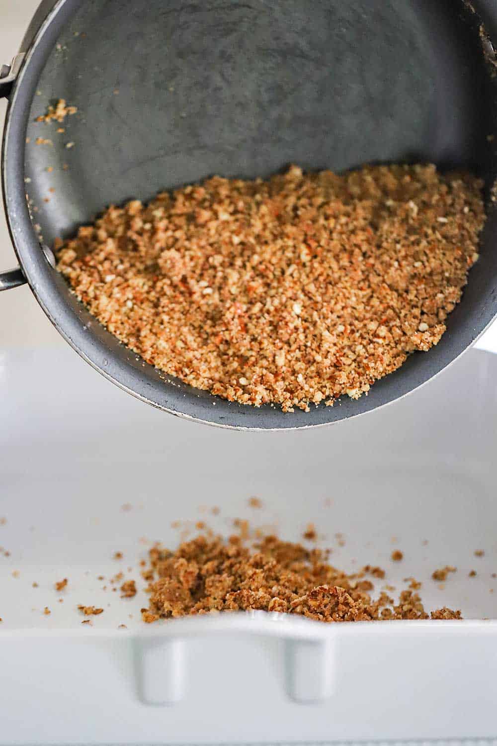 A crushed pretzel and butter mixture being poured out of a pan into a square white baking dish. 