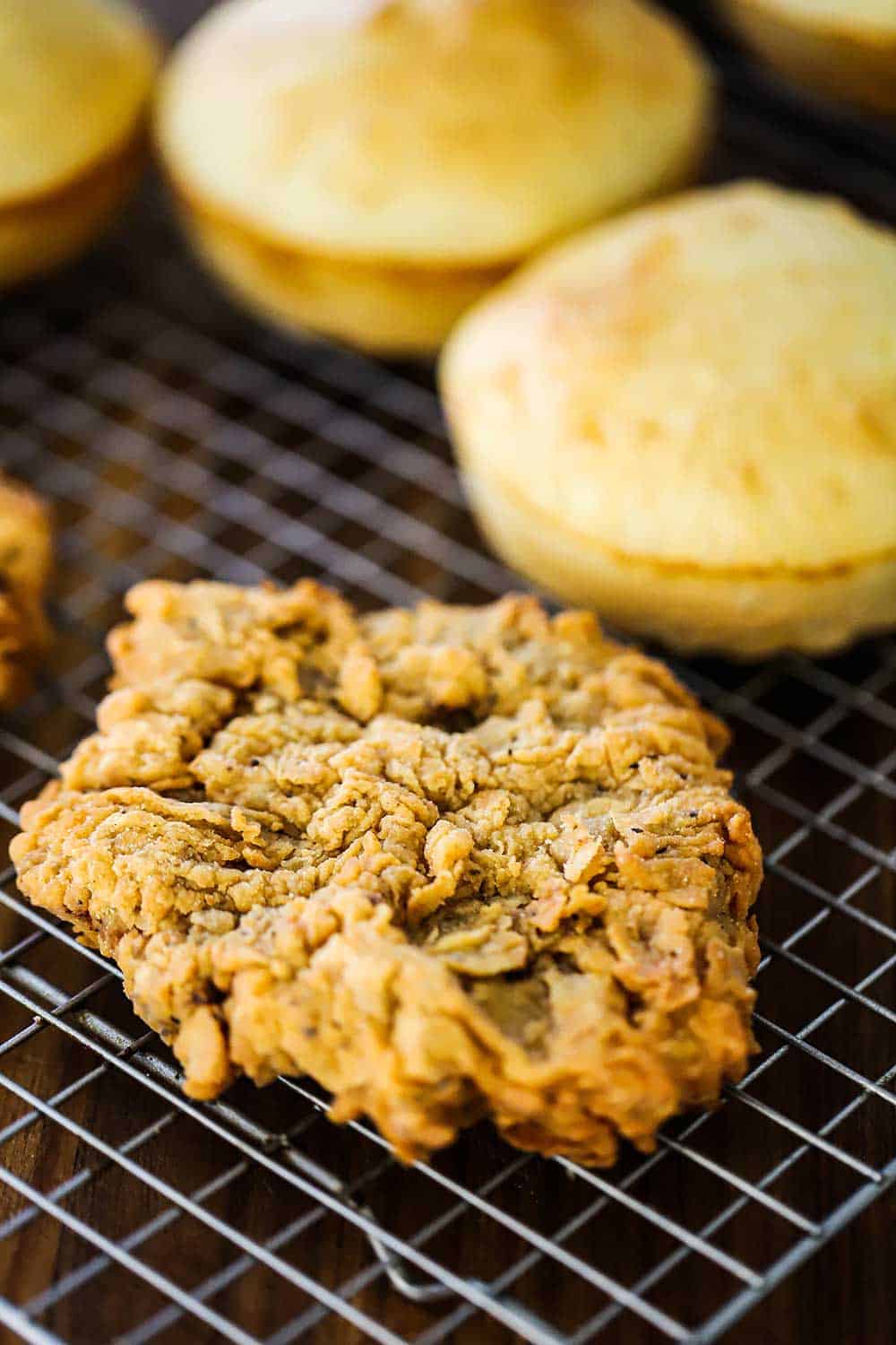 A chicken fried steak cutlet sitting on a baking rack next to homemade hamburger buns.