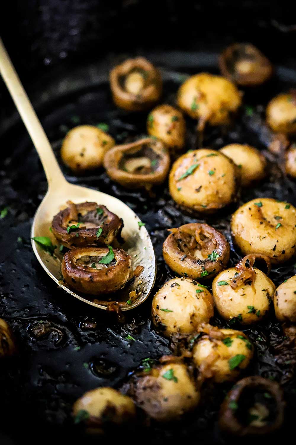 A black cast-iron skillet filled with sautéed mushrooms with a gold spoon holding a couple of the mushrooms.