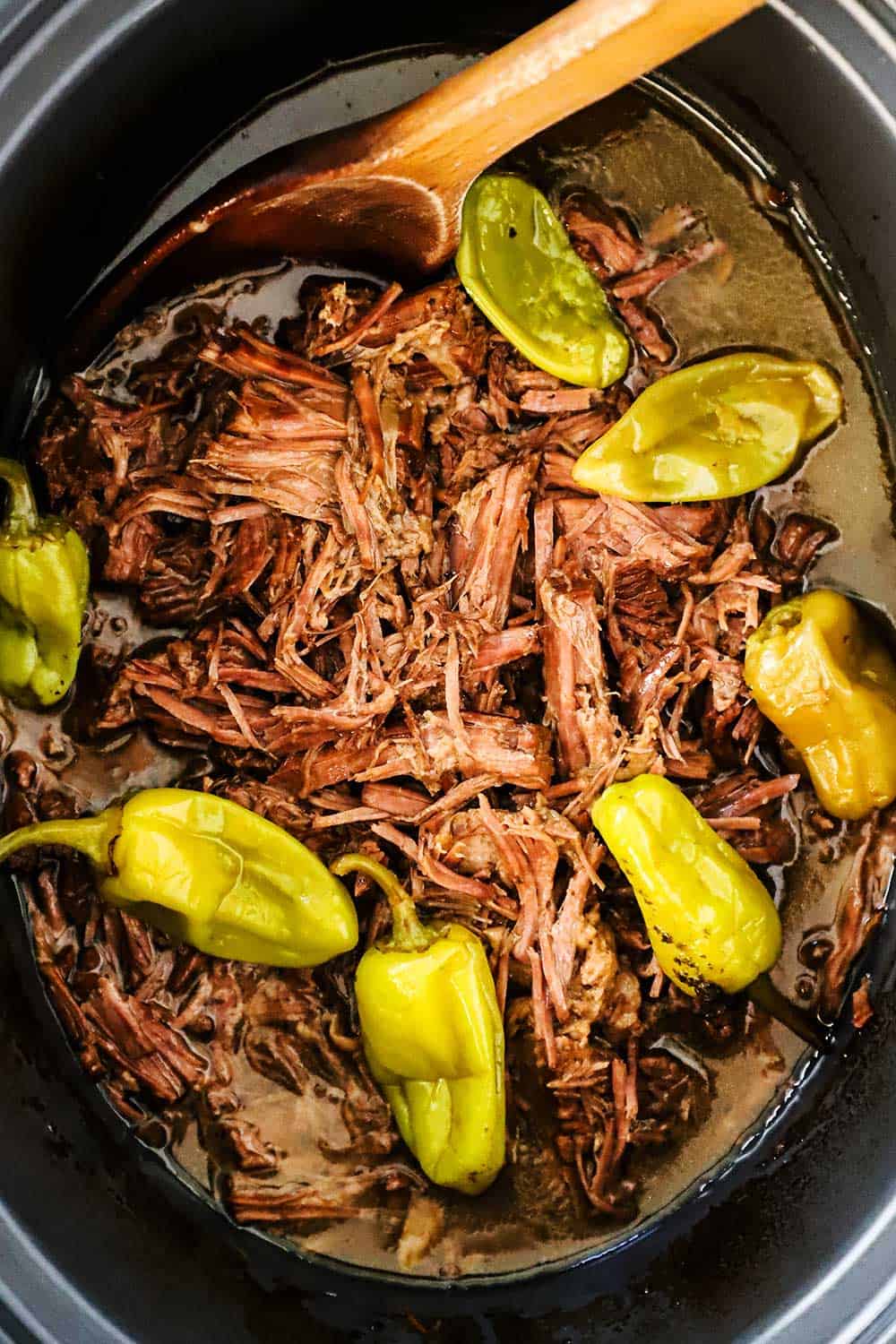 A slow-cooker filled with shredded pot roast topped with six pepperoncini peppers.