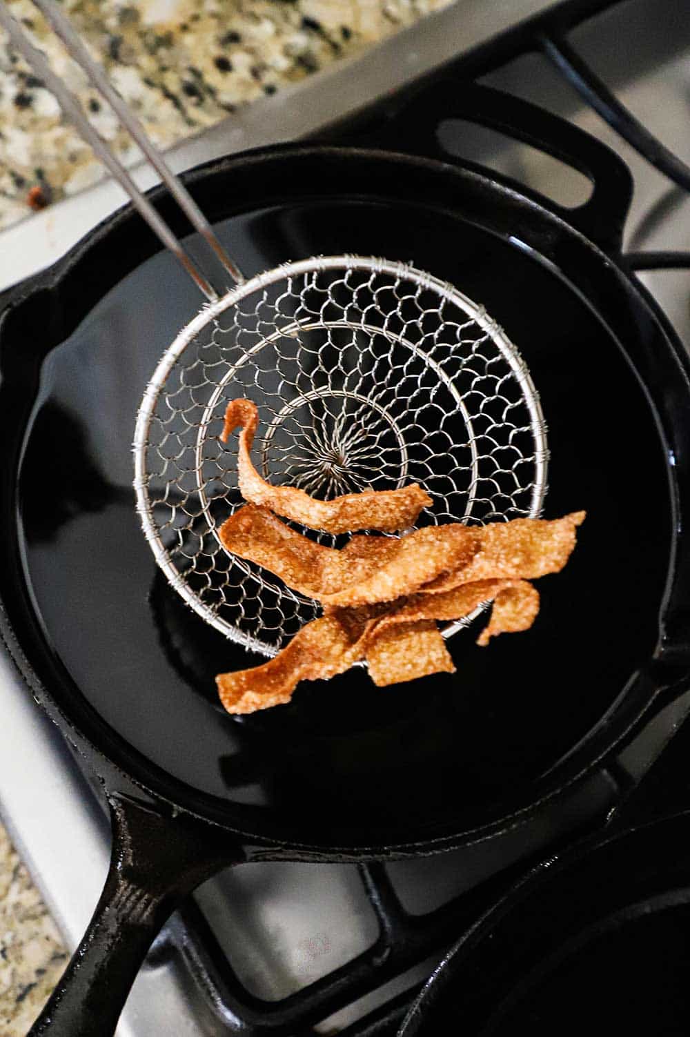 A metal spider lifting fried wonton strips out a cast-iron skillet filled with hot oil. 