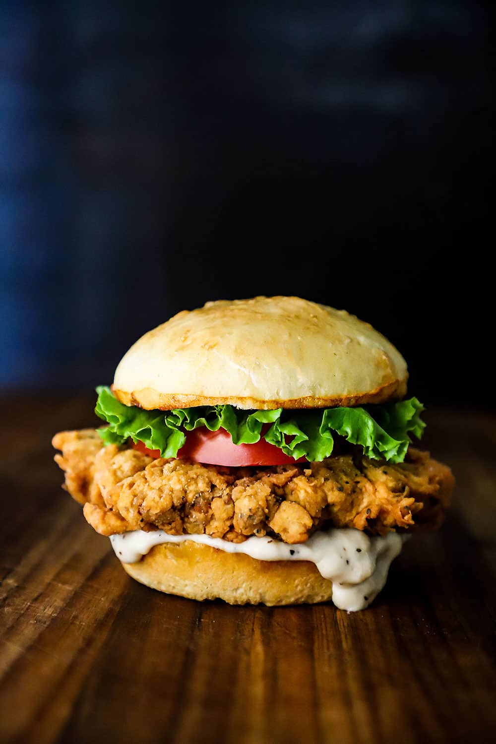 A chicken fried steak sandwich topped with sliced tomato and lettuce sitting on a wooden cutting board.