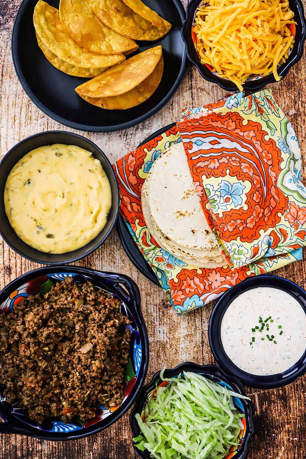 An assortment of bowls holding taco meat, shredded lettuce, spicy Ranch dressing, cheese sauce, taco shells, and shredded cheddar cheese.