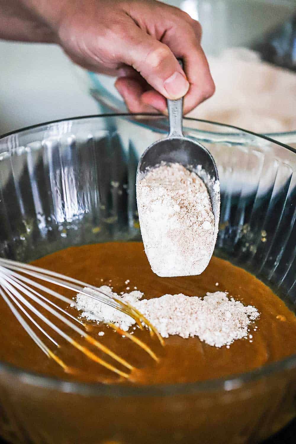 A person dumping flour from a metal scoop into a bowl filled with pumpkin batter.
