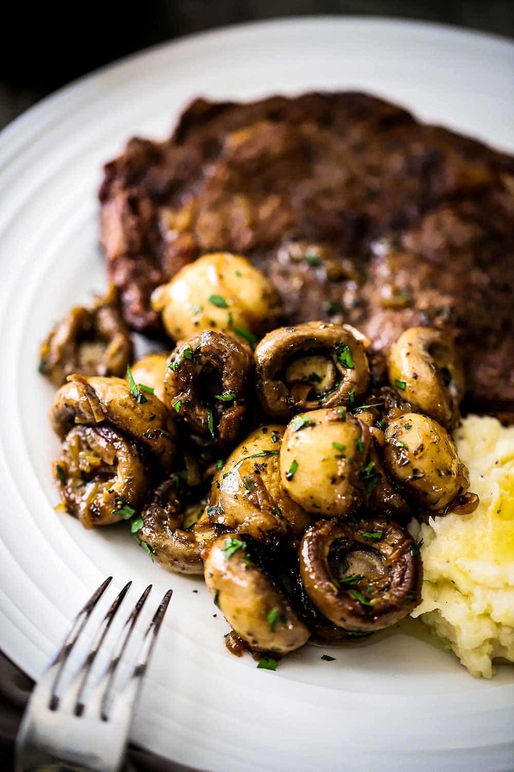 A white dinner plate filled with a grilled steak sitting next to a pile of sautéed whole mushrooms.
