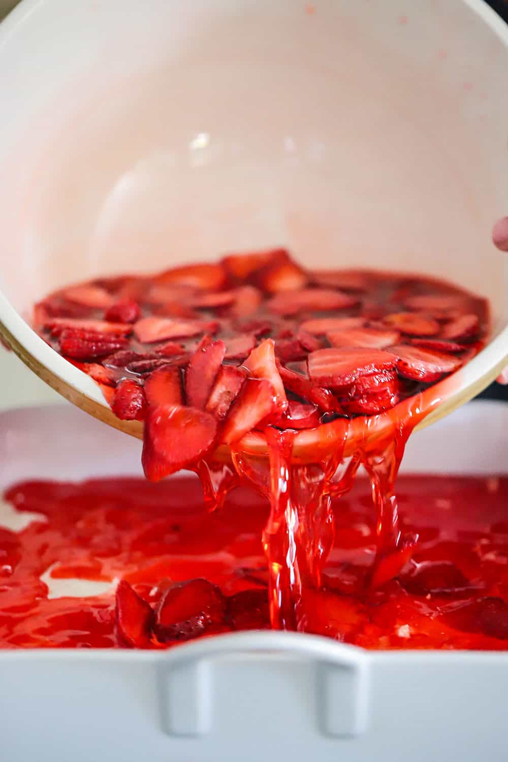 Strawberry Jell-O and sliced strawberries being poured from a ceramic bowl into a white baking dish for strawberry pretzel salad. 