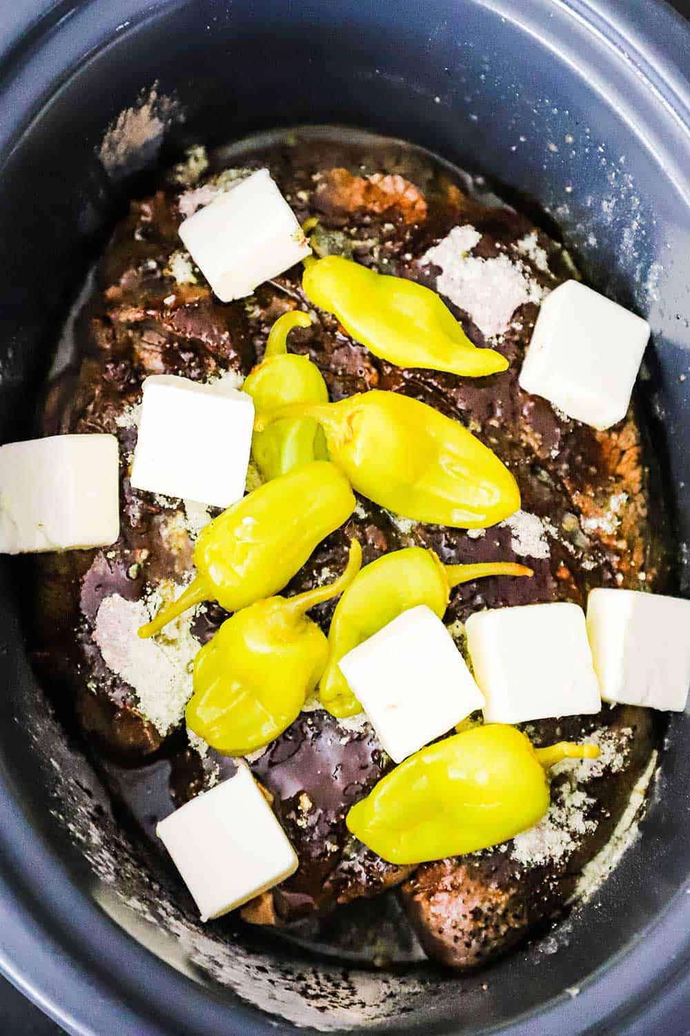 A chuck roast sitting in a slow-cooker topped with seasonings, peppers, and slices of chilled butter.