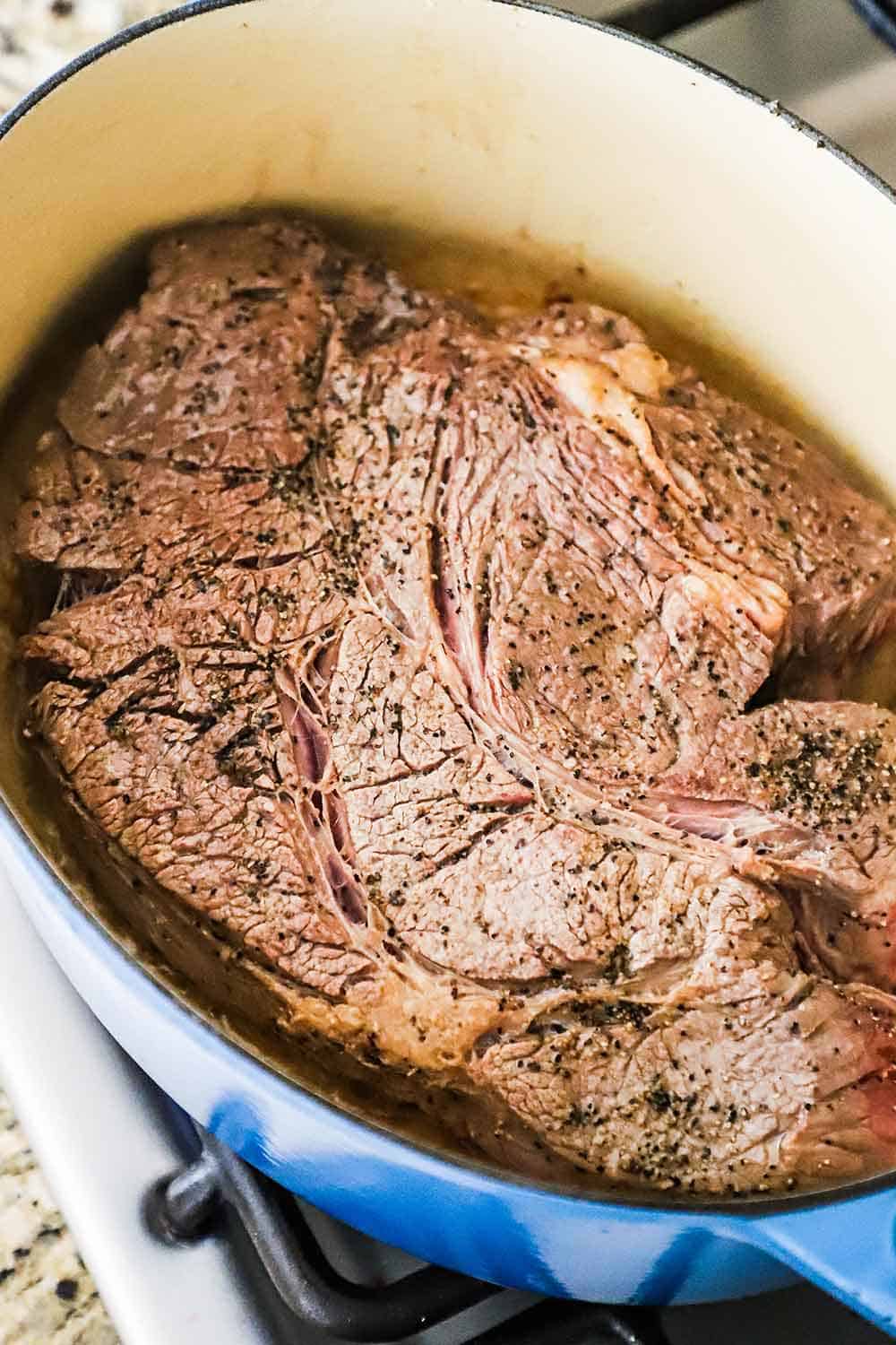 A large seared chuck roast sitting in a blue Dutch oven.