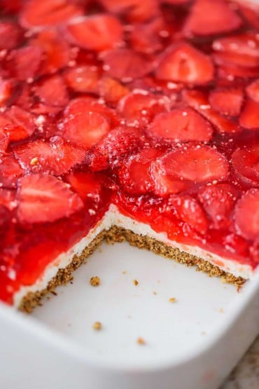 A square white casserole dish filled with strawberry pretzel salad with one piece missing in the corner.