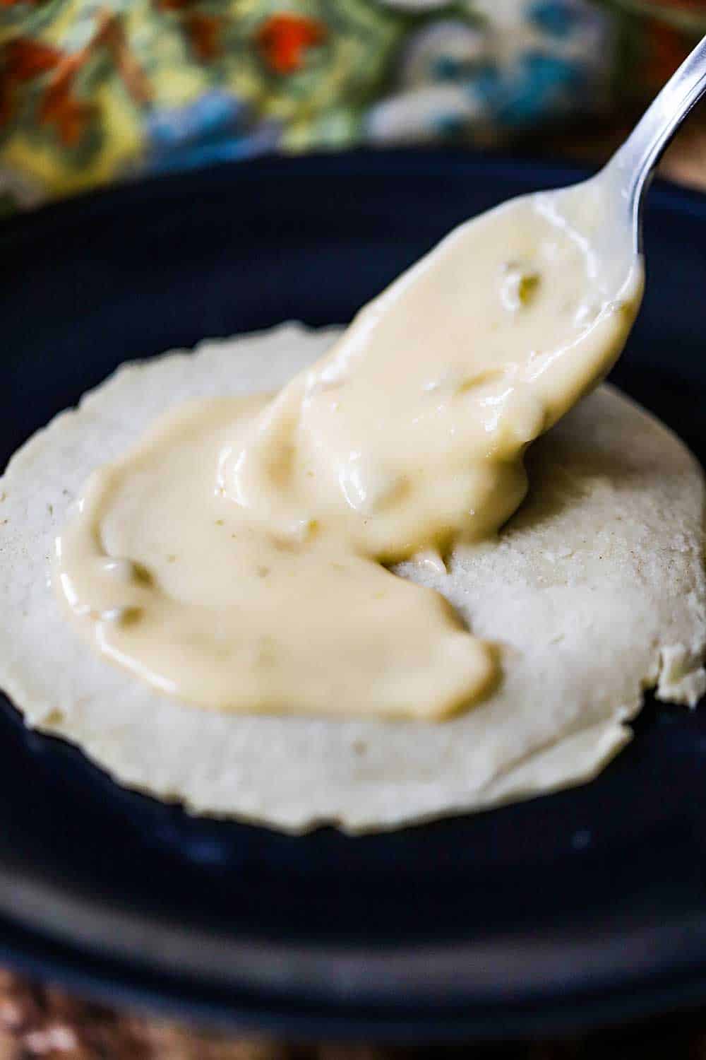 A large spoon spreading a cheese sauce over the surface of a homemade corn tortilla.