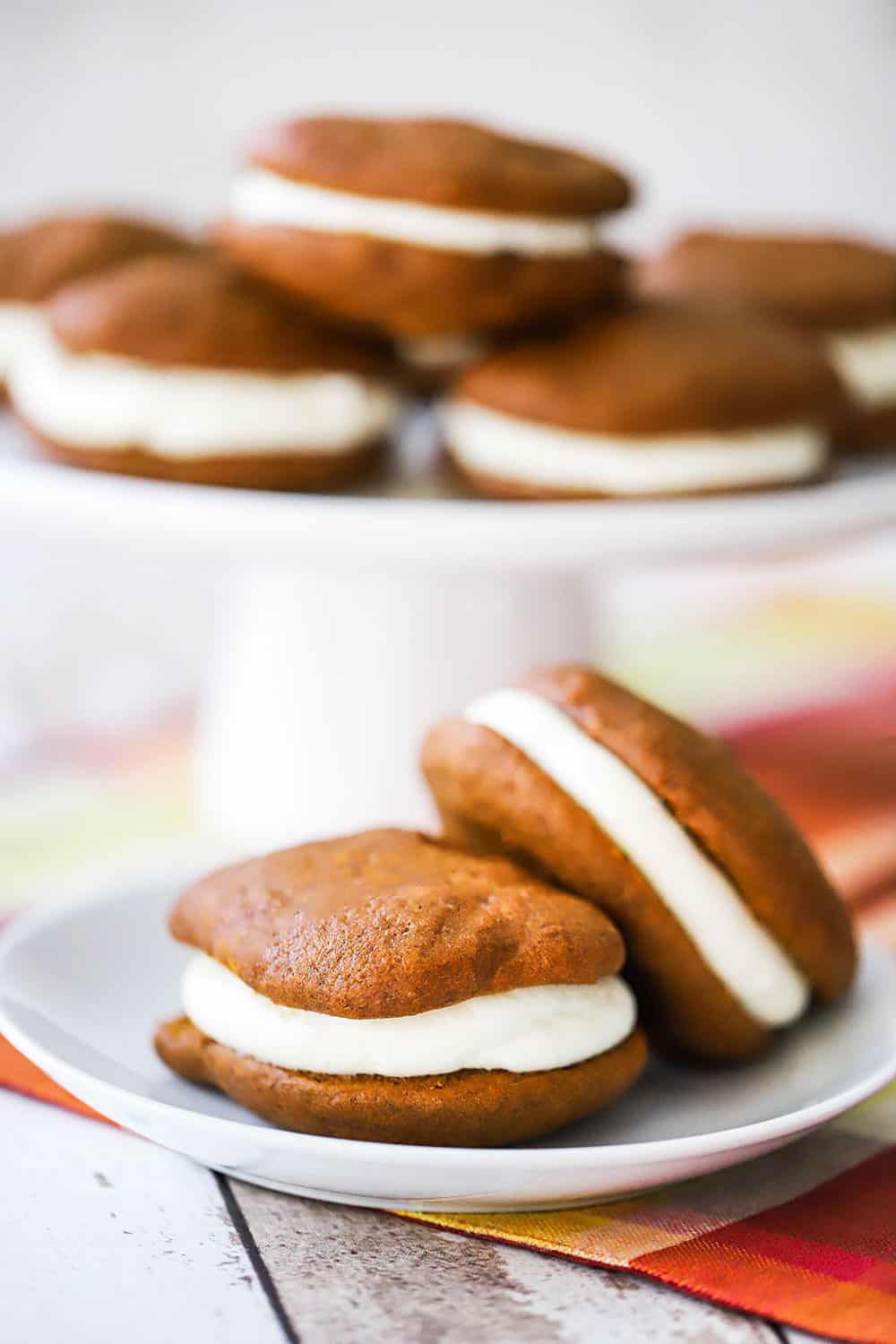 A white dessert plate holding two pumpkin whoopie pies in front of a cake platter holding more whoopie pies.