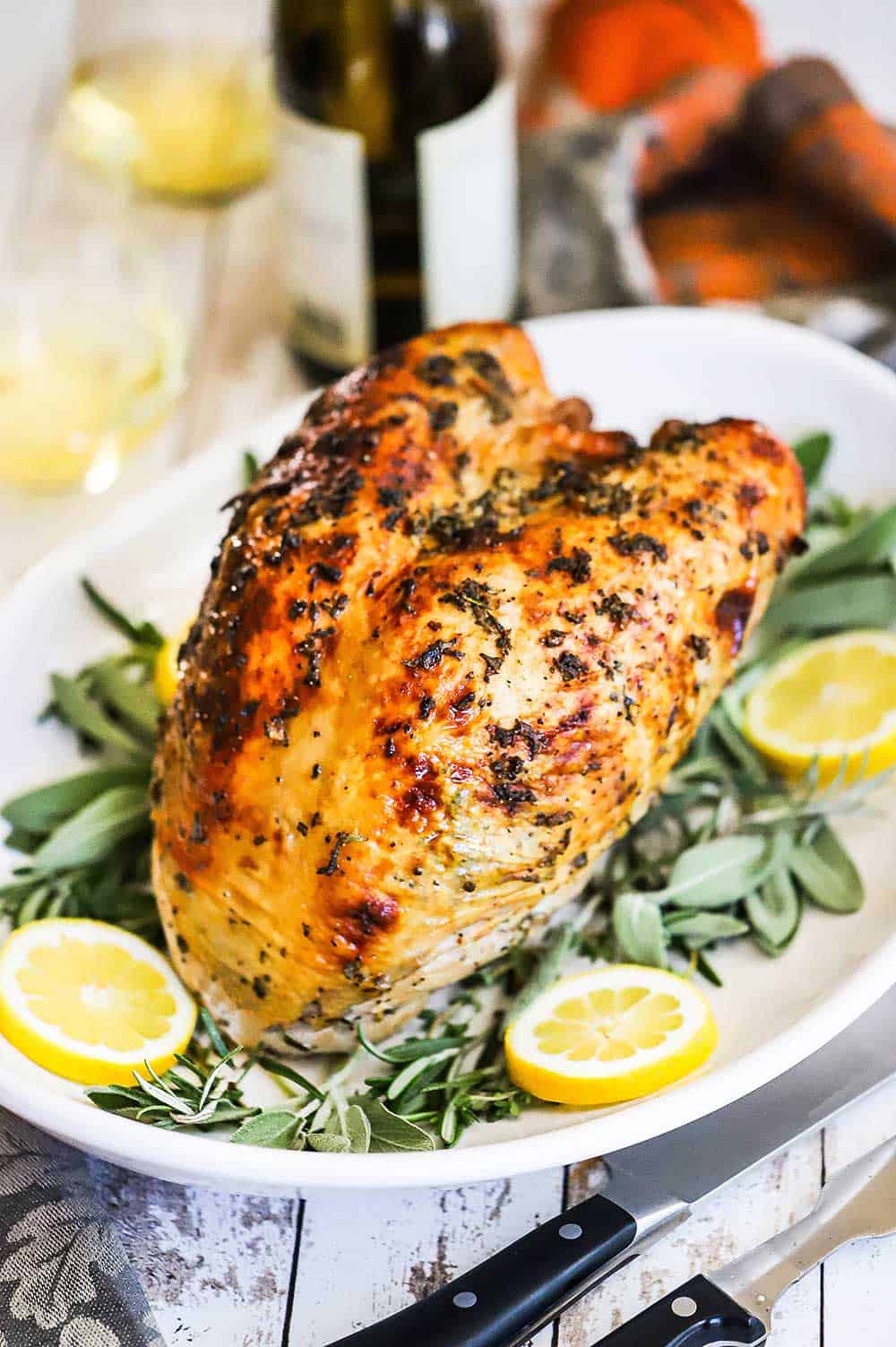 A roast turkey breast with herbs sitting on a platter lined with fresh herbs and lemon slices.