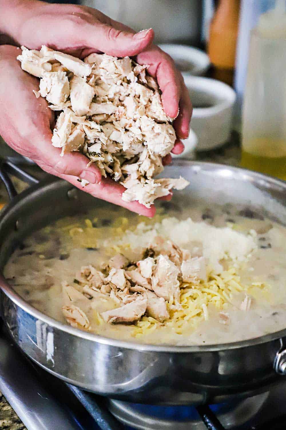 A pair of hands transferring chopped cooked turkey into a skillet filled with a sauce and shredded white cheddar cheese. 