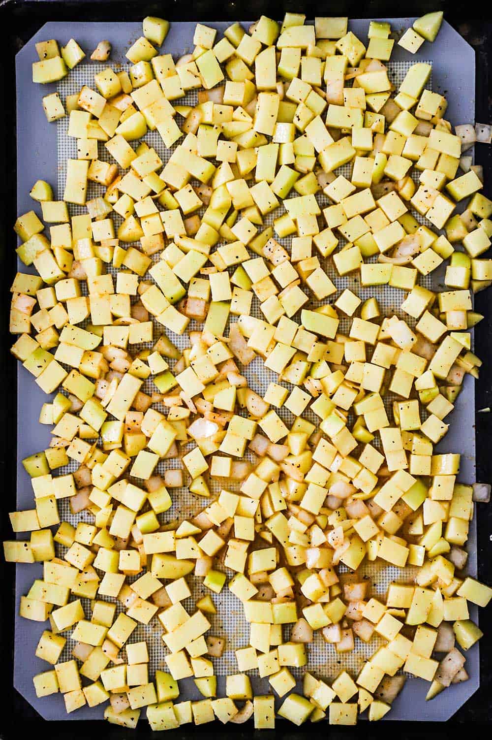 A rectangle baking pan lined with a silicone mat and topped with seasoned uncooked cubed potatoes. 