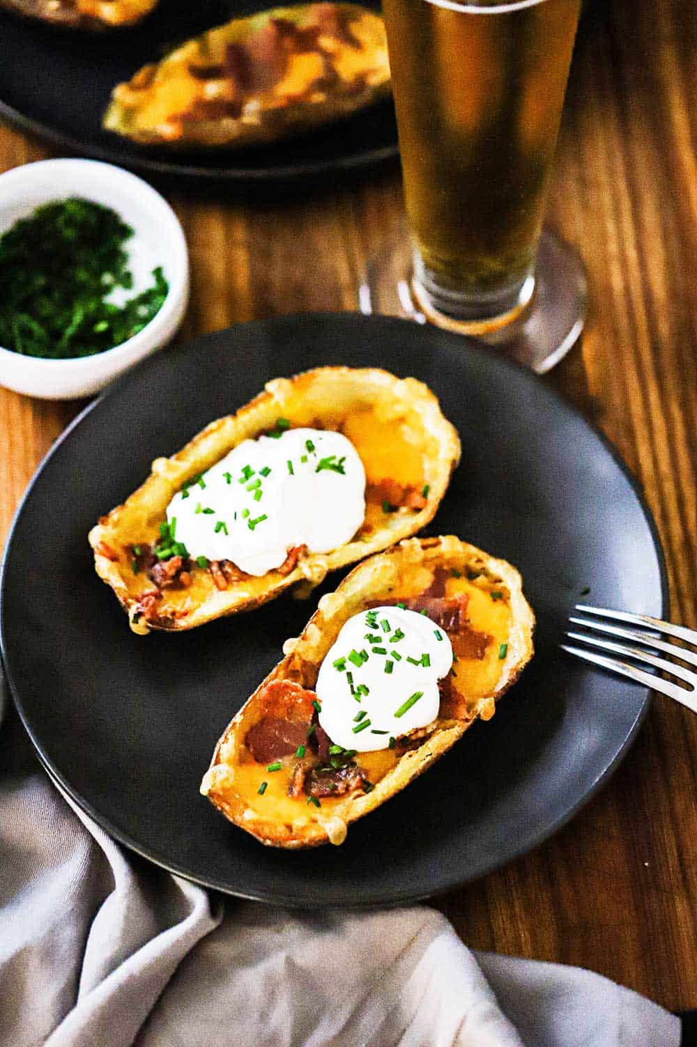 Two loaded potato skins sitting next to each other on a black plate with a tall beer next to it.