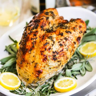 A roast turkey breast with herbs sitting on a platter lined with fresh herbs and lemon slices.
