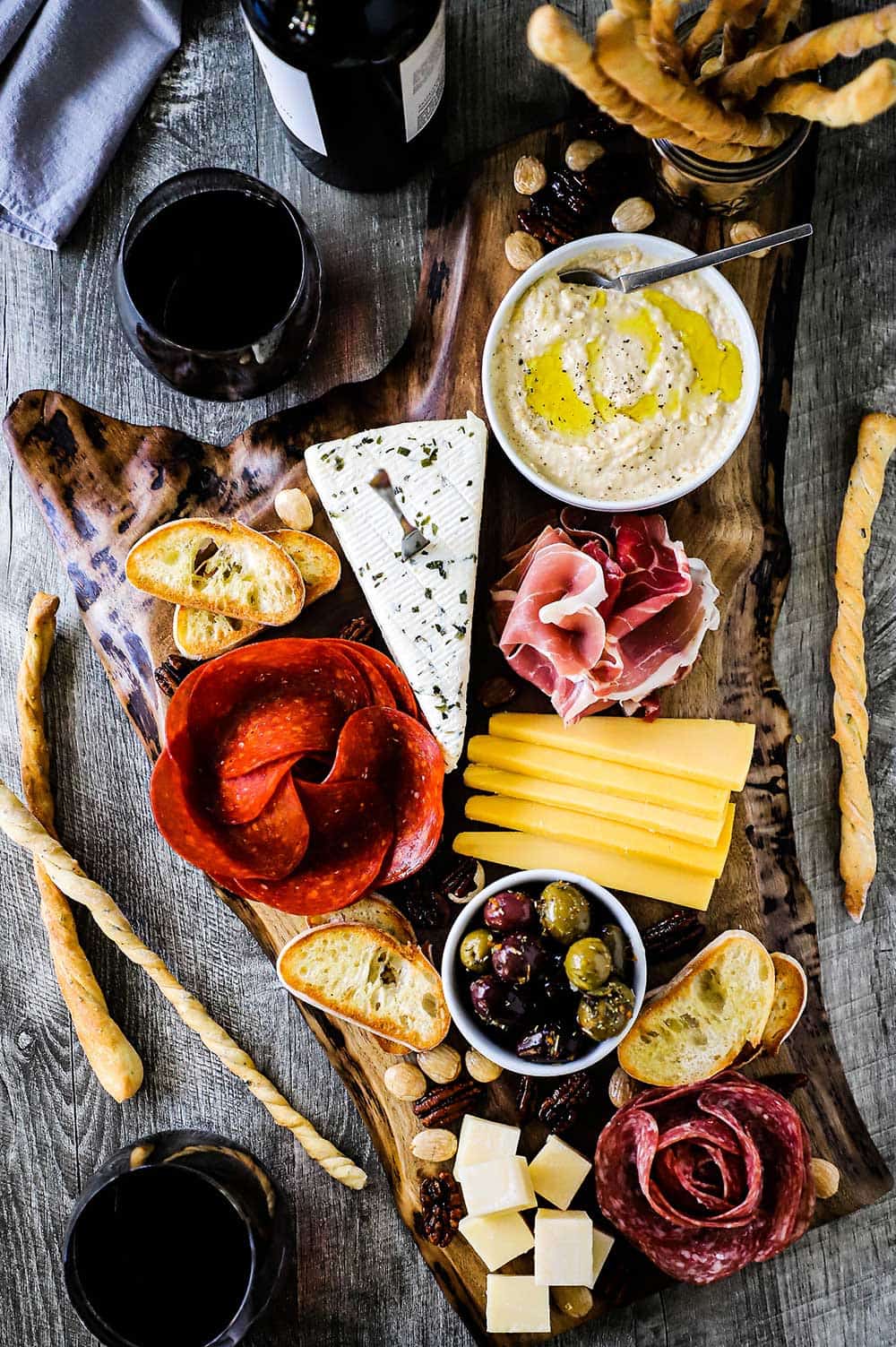 A charcuterie board that is covered with cubed cheese, a meat flower, hummus, olives, and bread sticks. 