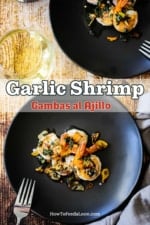 Two small dark grey appetizer plates filled with helpings of Spanish-style garlic shrimp both sitting next to glasses of white wine.