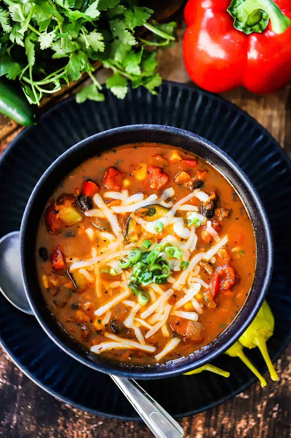 A blue soup bowl filled with vegetable chili and black beans next to a red bell pepper and a bunch of cilantro.