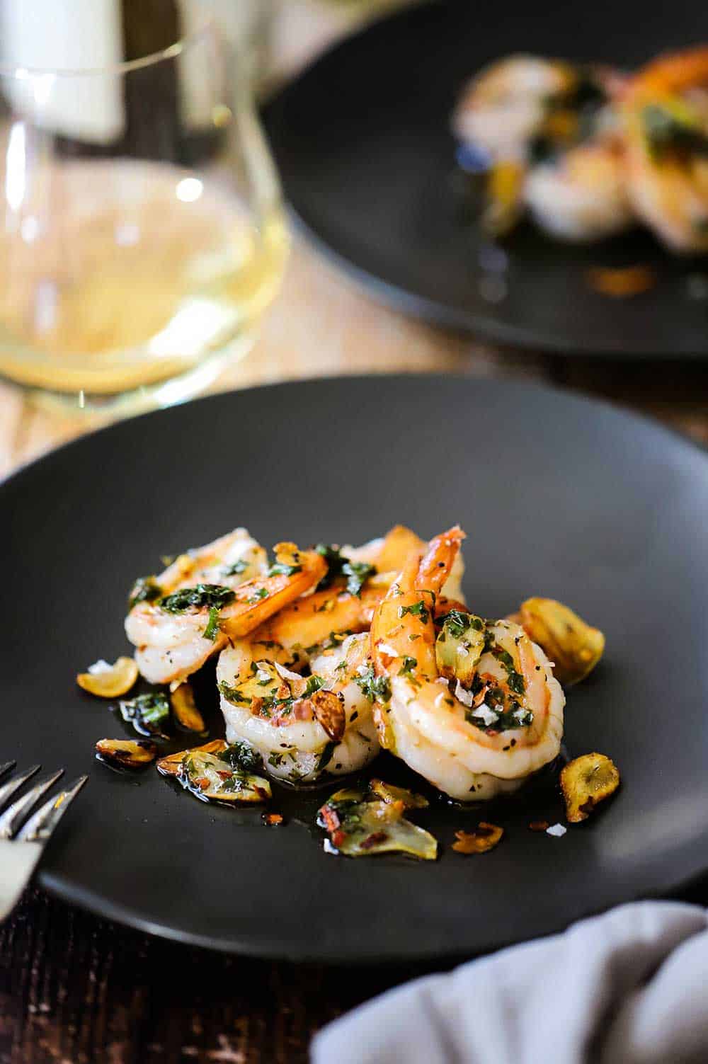 A small round dark grey plate filled with garlic shrimp sitting next to a stemless glass of white wine. 