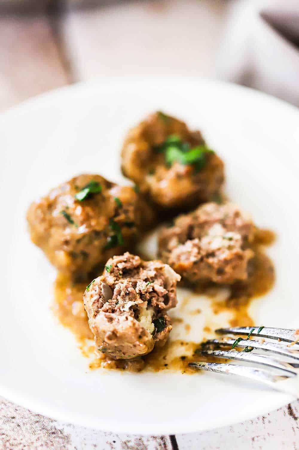 Three Swedish meatballs on a white cocktail plate with one of them broken open from a fork sitting next to the plate.