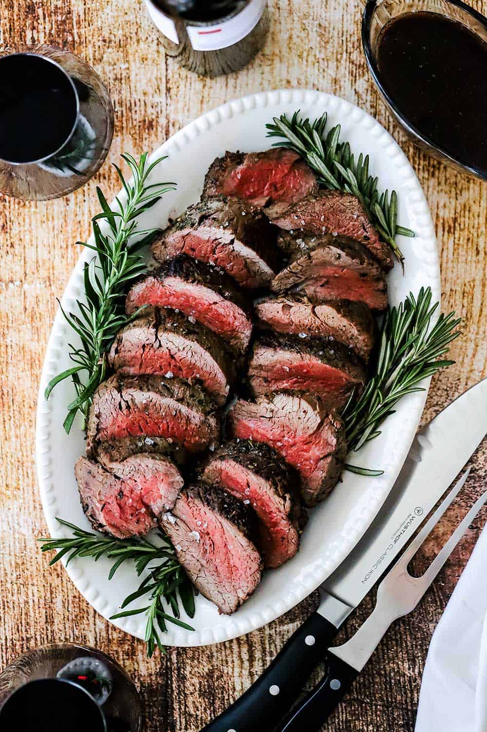 An oval white platter filled with thick slices of beef tenderloin surrounded by fresh rosemary and sitting next to a gravy boat filled with red wine sauce. 