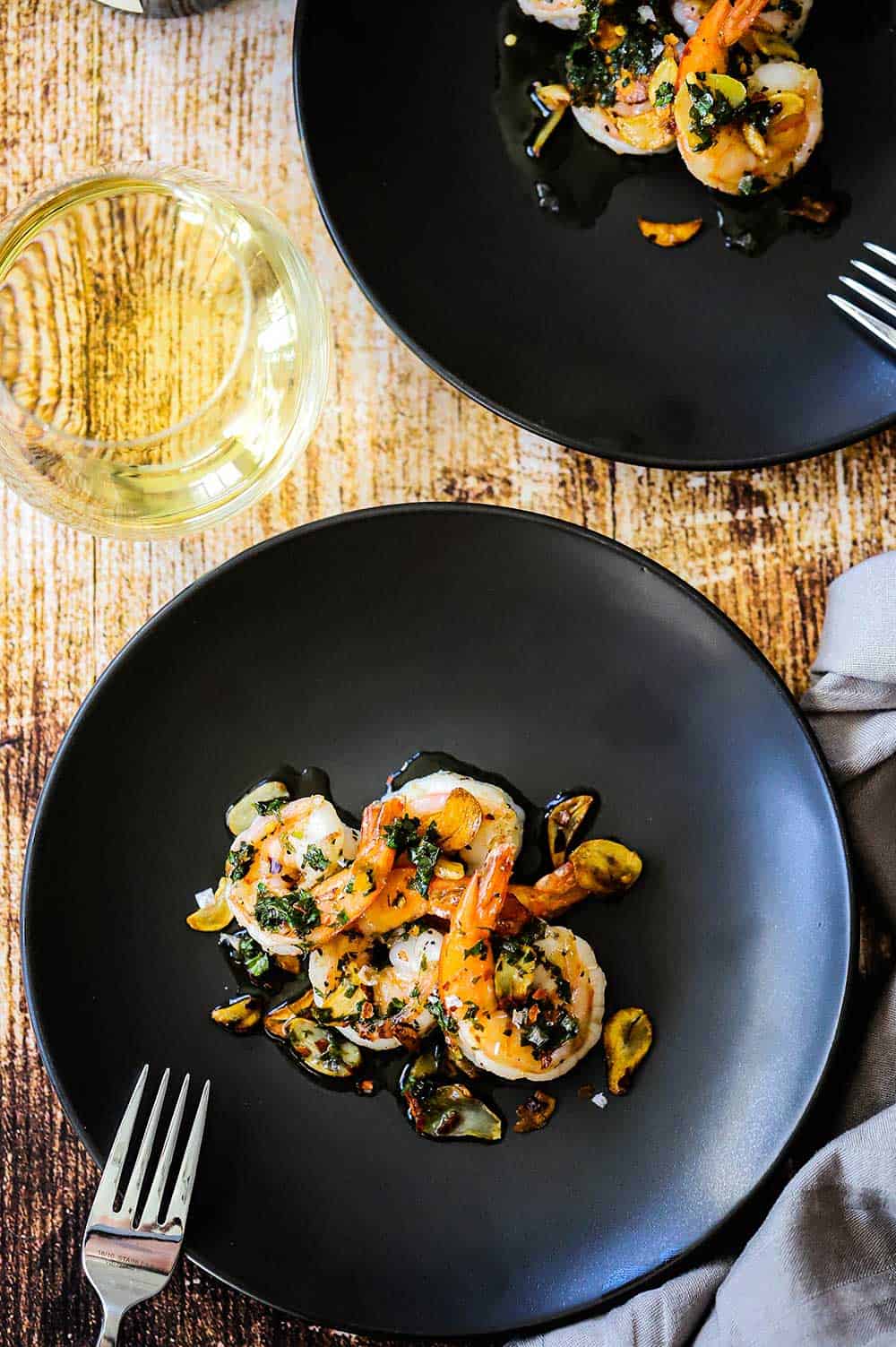 Two small dark grey appetizer plates filled with helpings of Spanish-style garlic shrimp both sitting next to glasses of white wine. 