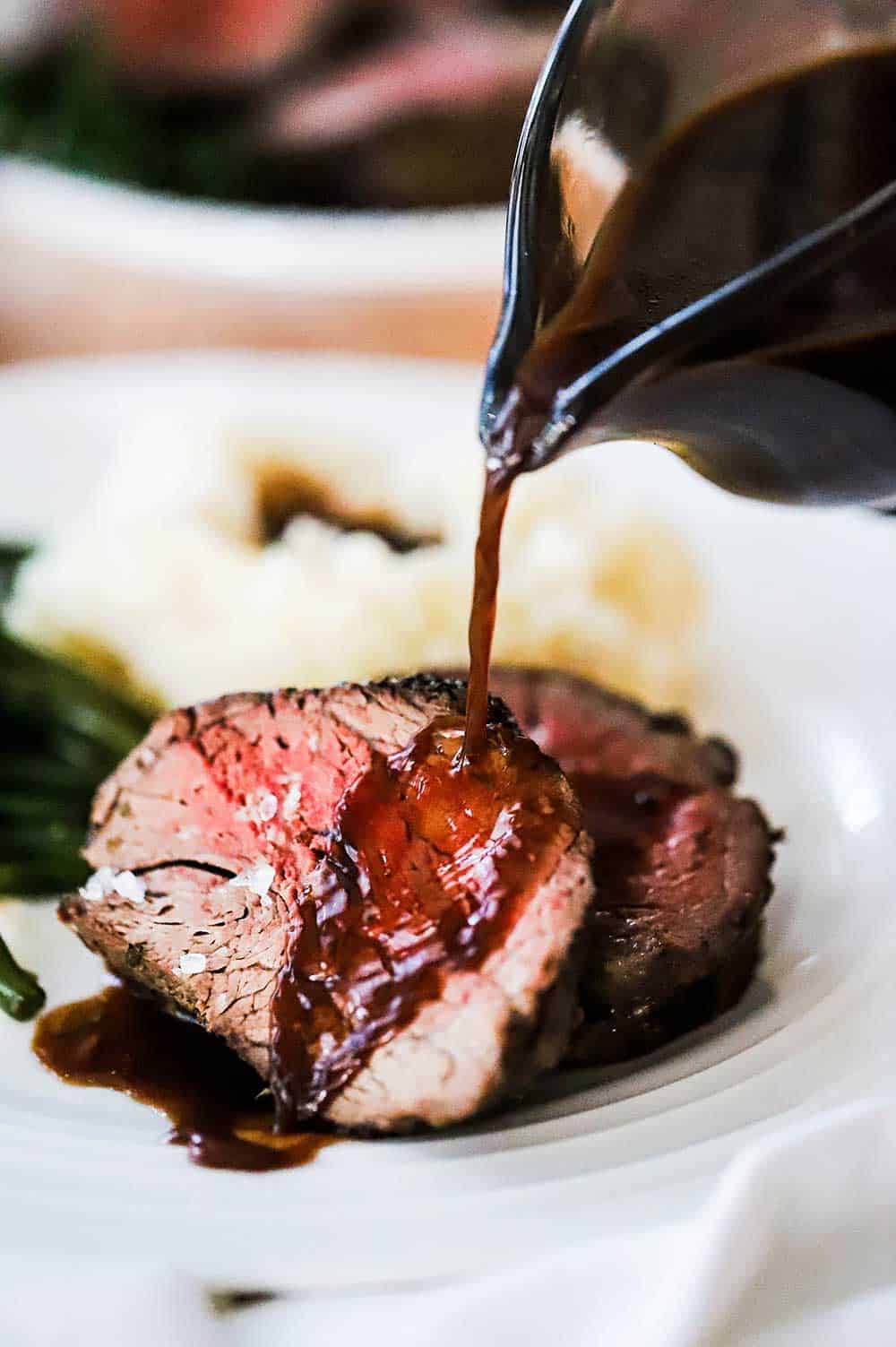 Red wine sauce being poured from a glass gravy boat onto two slices of beef tenderloin on a plate next to mashed potatoes and green beans. 