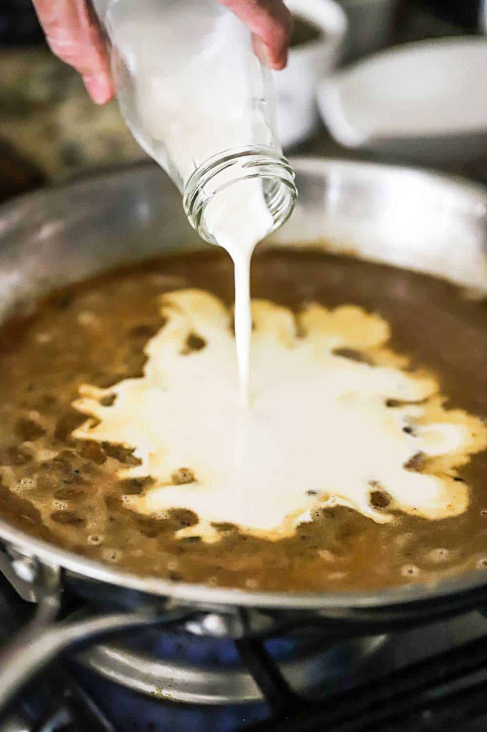 A person pouring heavy cream from a small milk jug into a skillet filled with a brown sauce.