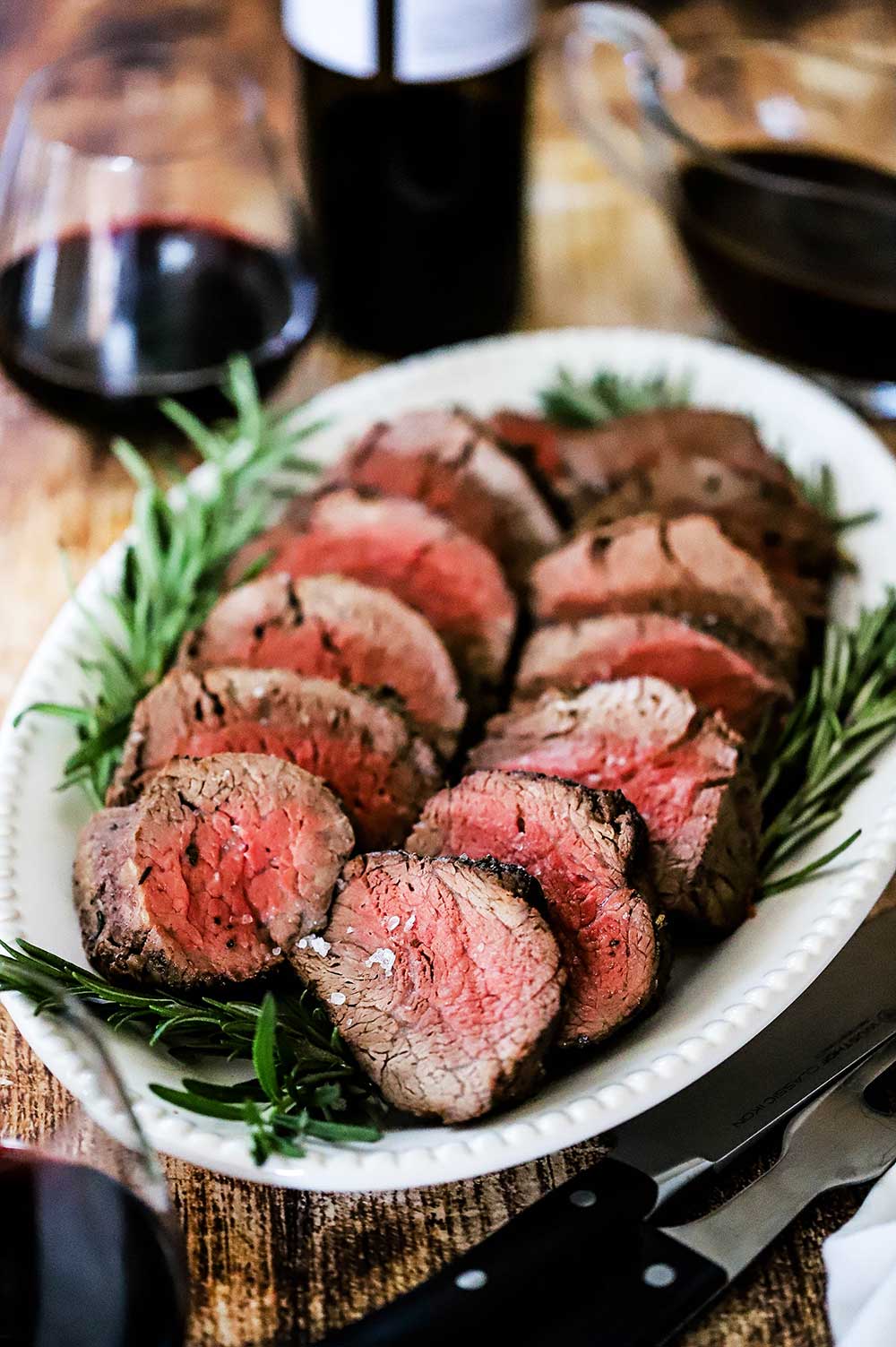 An oval white platter filled with slices of beef tenderloin with sprigs of rosemary along the edge and a glass or red wine sitting in the rear.